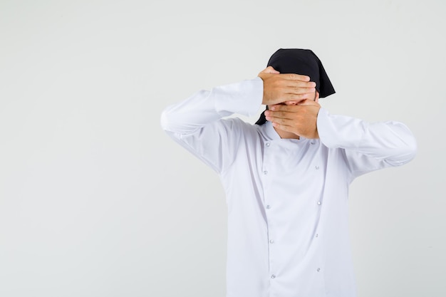 Chef masculino segurando a mão nos olhos e boca em uniforme branco e parecendo covarde. vista frontal.