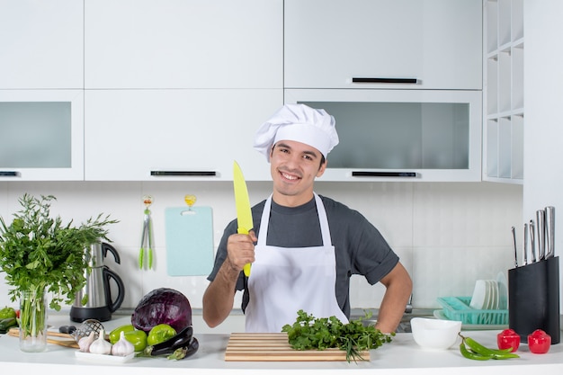 Chef masculino sorridente de frente e de uniforme segurando uma faca na cozinha