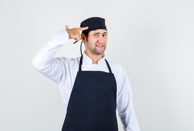 Chef masculino tocando a cabeça com gesto de arma de uniforme, vista frontal do avental.