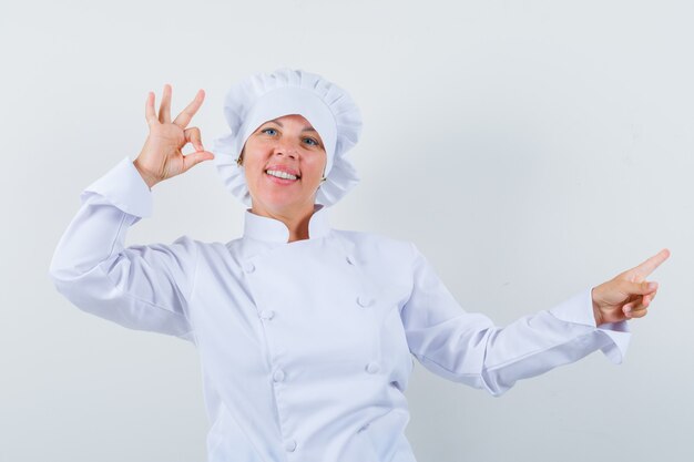 chef mulher apontando à parte, mostrando um gesto ok em uniforme branco e parecendo confiante.