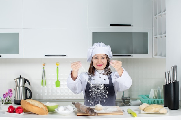 Chef positiva do commis feminino de uniforme em pé atrás da mesa, manchando o rosto com farinha na cozinha branca