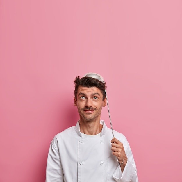 Chef positivo posa com a concha na cabeça, vai preparar a sopa, usa uniforme branco, segura os utensílios da cozinha, faz a cozinha do restaurante, olha para cima