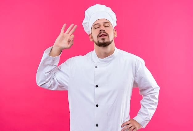 Chef profissional cozinheiro de uniforme branco e chapéu de cozinheiro, fazendo sinal de ok com os olhos fechados, em pé sobre um fundo rosa
