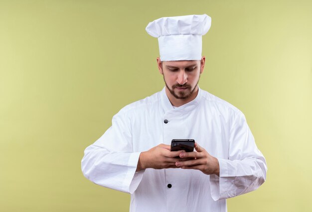 Chef profissional cozinheiro de uniforme branco e chapéu de cozinheiro segurando um smartphone, conversando com alguém em pé sobre um fundo verde