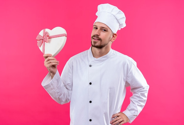 Chef profissional cozinheiro de uniforme branco e chapéu de cozinheiro segurando uma caixa de presente, parecendo confiante sorrindo sobre o fundo rosa