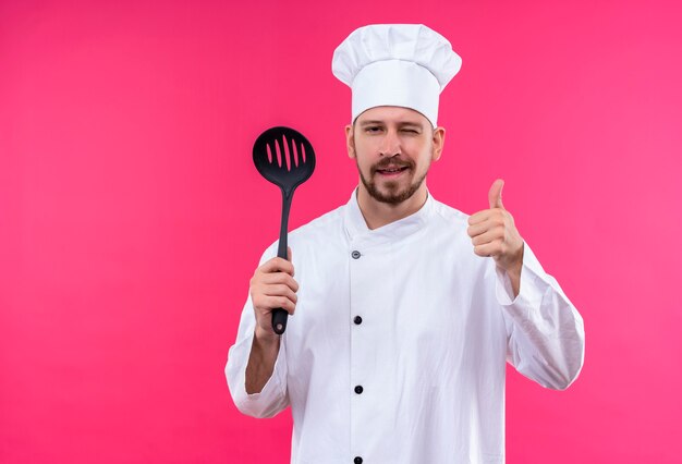 Chef profissional cozinheiro de uniforme branco e chapéu de cozinheiro segurando uma concha, olhando para a câmera, piscando e sorrindo, mostrando os polegares em pé sobre o fundo rosa