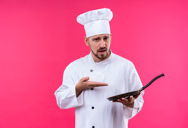 Chef profissional cozinheiro de uniforme branco e chapéu de cozinheiro segurando uma panela apontando para ela com o braço da mão, olhando para a câmera com expressão cética no rosto em pé sobre um fundo rosa