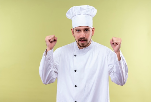 Chef profissional cozinheiro em uniforme branco e chapéu de cozinheiro cerrando os punhos, feliz e animado, regozijando-se com seu sucesso em pé sobre fundo verde