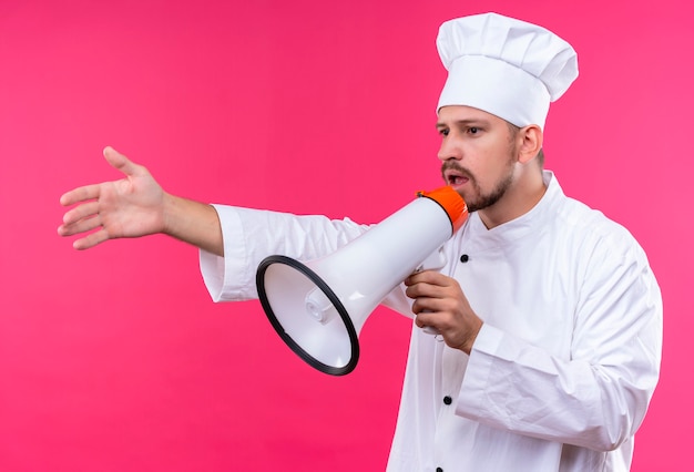 Chef profissional cozinheiro em uniforme branco e chapéu de cozinheiro falando para o megafone gesticulando com a mão em pé sobre o fundo rosa