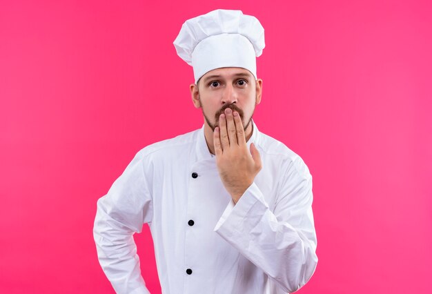 Chef profissional cozinheiro em uniforme branco e chapéu de cozinheiro parecendo espantado e surpreso, cobrindo a boca com a mão em pé sobre um fundo rosa