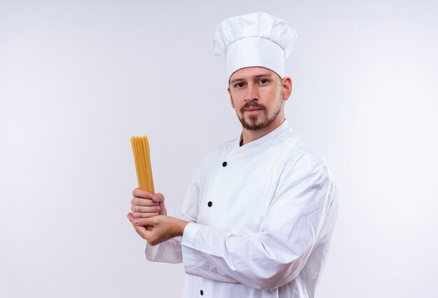 Chef profissional cozinheiro em uniforme branco e chapéu de cozinheiro segurando macarrão espaguete cru, parecendo confiante em pé sobre um fundo branco