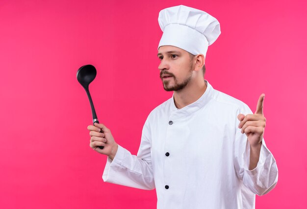 Chef profissional cozinheiro em uniforme branco e chapéu de cozinheiro segurando uma concha apontando o dedo para cima, tendo uma ótima ideia em pé sobre um fundo rosa