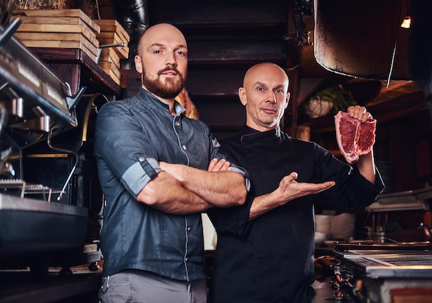 Chef segurando um bife fresco e seu assistente de pé perto com os braços cruzados em uma cozinha de restaurante.