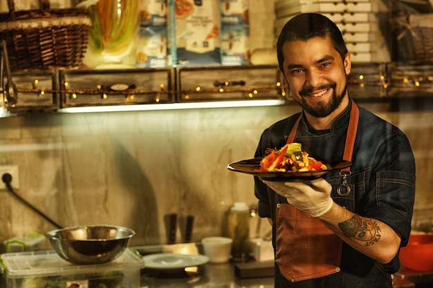 Foto grátis chef sorrindo e mostrando deliciosa salada feita de legumes frescos homem segurando prato com salada em sua mão fundo de cozinha de restaurante profissional com utensílios de cozinha especiais