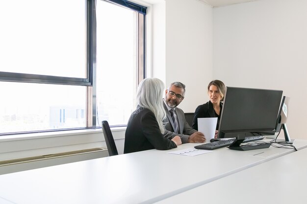 Chefe e gerentes analisando relatórios e discutindo o trabalho. Equipe sentados juntos no local de trabalho com monitores, papéis e conversando. Copie o espaço. Conceito de reunião de negócios