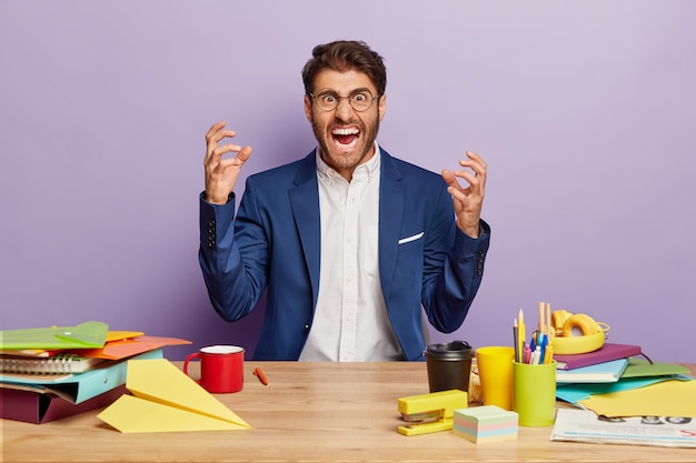 Foto grátis chefe masculino irritado usa terno elegante, levanta a mão e grita com raiva para os colegas, exige trabalhar a tempo, senta-se à mesa de madeira com café para viagem