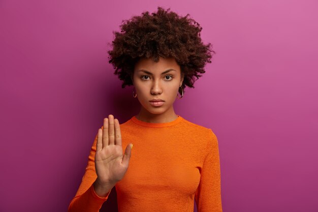 Chega, por favor. Mulher séria e rígida faz gesto de pare, mostra proibição e pede espera, recusa algo, veste suéter laranja, isolado na parede roxa. Não significa nunca, não para isso