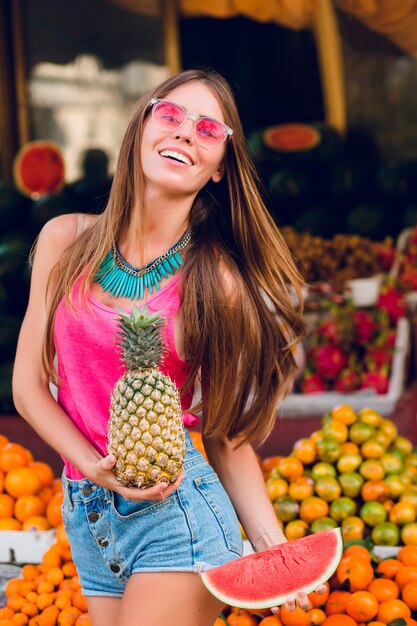 Cheio de alegria garota verão se divertindo no mercado de frutas tropicais. Ela segura ananas, fatia de melancia e sorrindo