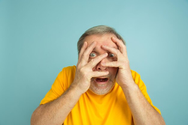 Chocado, cobrindo o rosto com as mãos. Retrato de homem caucasiano sobre fundo azul do estúdio. Lindo modelo masculino posando de camisa amarela.