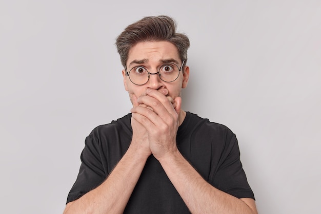 Chocado e mudo europeu jovem cobre a boca com as mãos gravatas para ser mudo parece com medo na câmera usa óculos e poses de camisa casual t contra um fundo branco. manter o conceito de silêncio