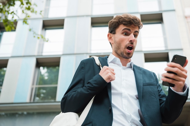 Chocado jovem empresário com sua mochila, olhando para o celular