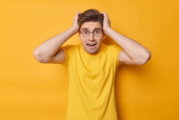 Chocado preocupado adulto homem europeu de cabelos escuros agarra a cabeça pensa no prazo parece estressado usa camiseta casual isolada sobre fundo amarelo Pessoas reações humanas e conceito de emoções