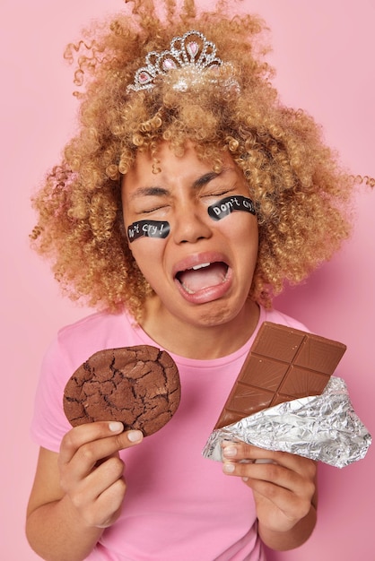 Foto grátis chorando mulher chateada com cabelo encaracolado usa coroa pequena tem vício em açúcar segura biscoito e barra de chocolate tem expressão triste isolada sobre fundo rosa estresse após quebrar a dieta