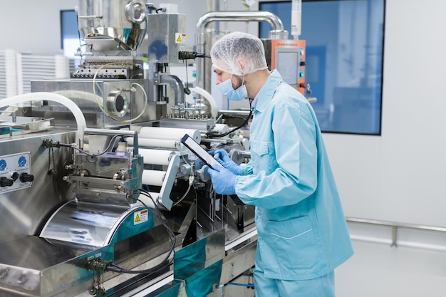 Cientista caucasiano em uniforme de laboratório azul configura máquina de fabricação com eixos e painel de controle verificando leituras fazendo anotações