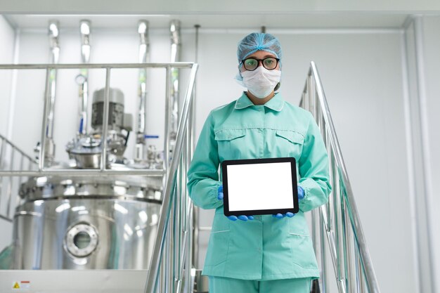 Cientista caucasiano em uniforme de laboratório azul e óculos ficam em escadas cromadas e seguram tablet vazio em direção à câmera