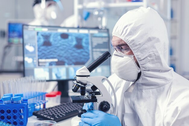 Cientista concentrado em equipamento ppe olhando para o microscópio em laboratório. Cientista em traje de proteção, sentado no local de trabalho, usando tecnologia médica moderna durante a epidemia global.