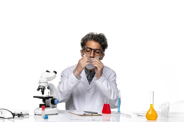 Cientista de meia-idade com vista frontal em um terno branco especial sentado ao redor da mesa com soluções