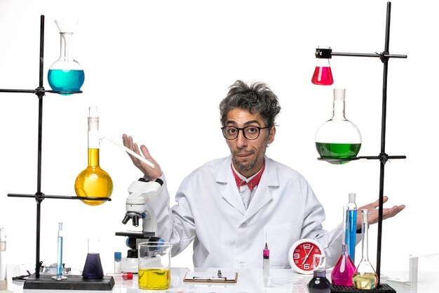 Cientista masculino de frente para o hospital em terno médico sentado em frente à mesa com soluções em saúde de vírus de laboratório cobiçado química de fundo branco
