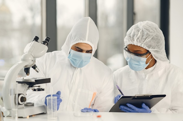 Cientistas e microbiologistas com traje de EPI e máscara facial seguram tubo de ensaio e microscópio em laboratório, encontrando tratamento ou vacina para infecção por coronavírus. Covid-19, laboratório e conceito de vacina.