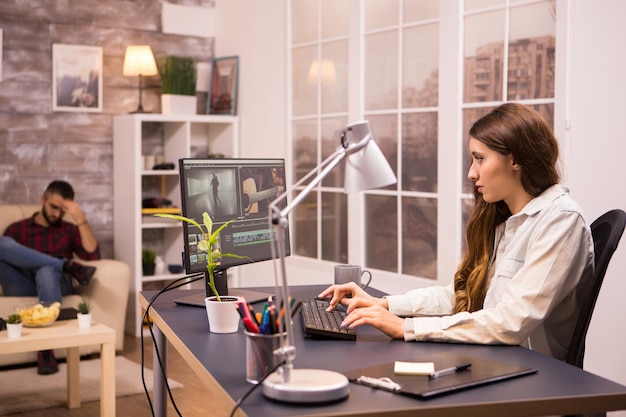 Cinegrafista feminina criativa trabalhando em um filme usando seu computador doméstico. Namorado relaxando no sofá ao fundo.