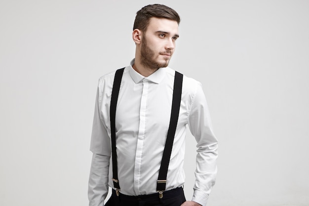 Cintura para cima foto de cara bonito na moda com barba elegante em pé na parede do estúdio branco com um olhar positivo de um sonho, vestido com uma camisa branca elegante com suspensórios. Conceito de pessoas e estilo de vida