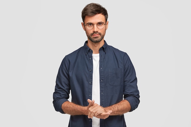 Cintura para cima, retrato de bonito homem sério com barba por fazer mantém as mãos juntas, vestido com camisa azul escura, tem conversa com o interlocutor, fica contra a parede branca. Freelancer homem autoconfiante