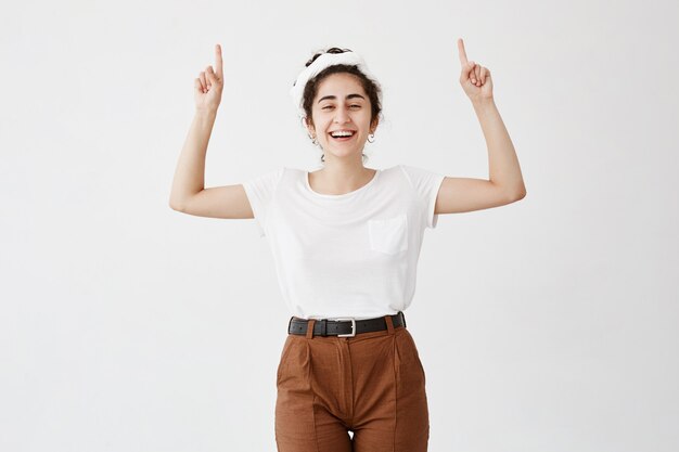 Cintura para cima tiro de alegre garota feliz vestindo camiseta branca, apontando os dedos no espaço da cópia acima de sua cabeça. Jovem mulher indicando algo na parede em branco com as mãos