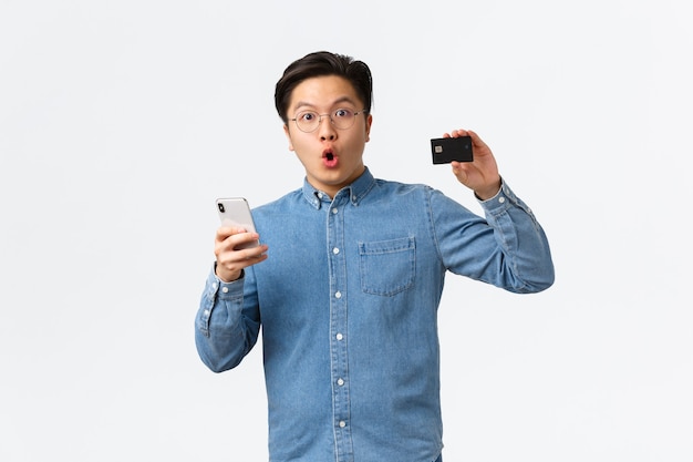 Cliente de banco asiático impressionado e animado, cara de óculos e roupas casuais mostrando cartão de crédito enquanto usa o smartphone para abrir o aplicativo de e-banking, fazendo compras na internet com o aplicativo móvel.