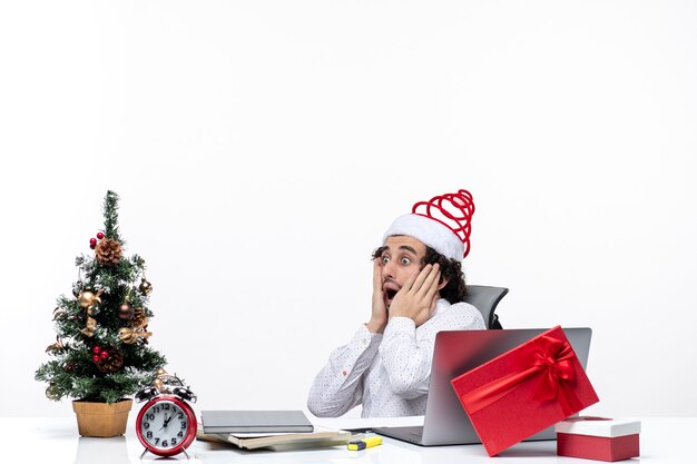 Clima de ano novo com jovem empresário chocado e animado com chapéu de Papai Noel sentado no escritório sobre fundo branco