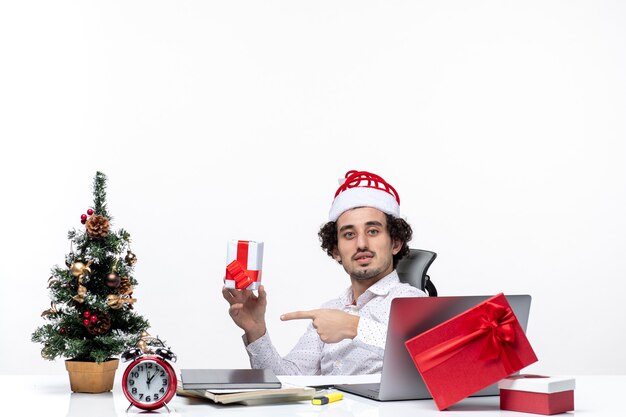 Clima de ano novo com jovem empresário com chapéu de Papai Noel sentado no escritório e apontando seu presente, posando para a câmera em fundo branco