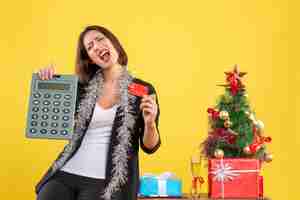 Foto grátis clima de natal com a bela moça emocional em pé no escritório e segurando o cartão do banco da calculadora no escritório em amarelo