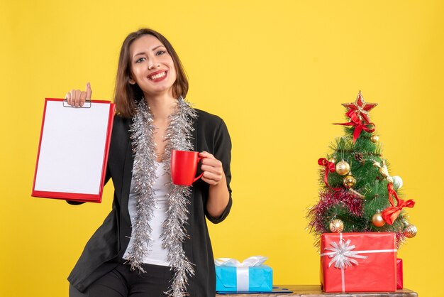 Clima de Natal com a bela senhora sorridente em pé no escritório e segurando a xícara de documentos no escritório em amarelo