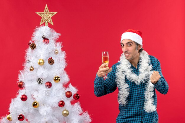 clima de natal com jovem engraçado emocional com chapéu de Papai Noel em uma camisa azul listrada segurando uma taça de vinho olhando para cima perto da árvore de Natal