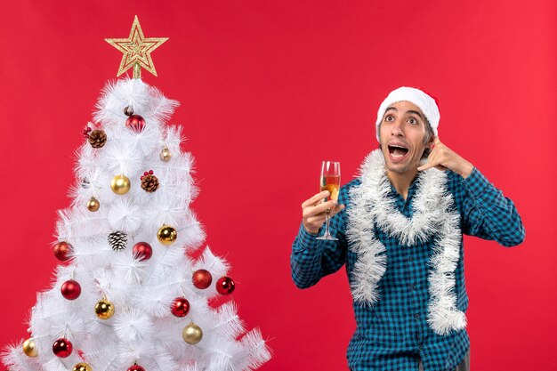 Clima de natal com jovem feliz com chapéu de papai noel em uma camisa azul listrada levantando uma taça de vinho e fazendo um gesto de me ligar perto da árvore de natal