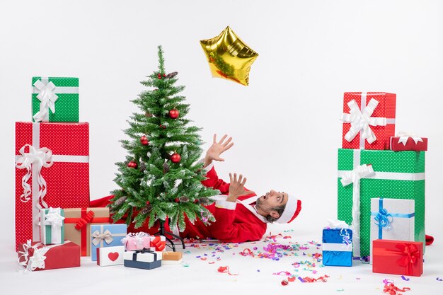 Clima de Natal com o jovem papai noel deitado atrás de uma árvore de natal perto de presentes em cores diferentes.