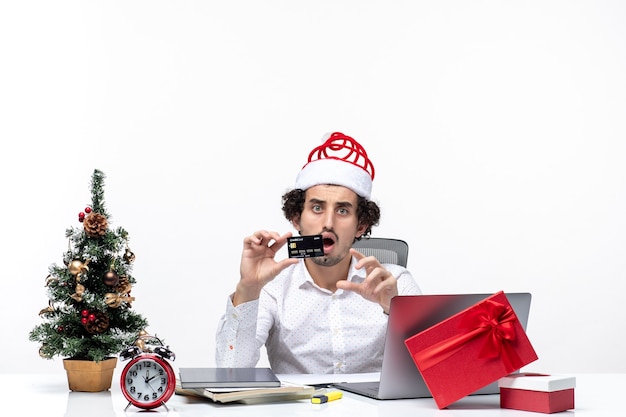 Clima festivo de feriado com empresário chocado com chapéu de Papai Noel e segurando o cartão do banco no escritório em fundo branco