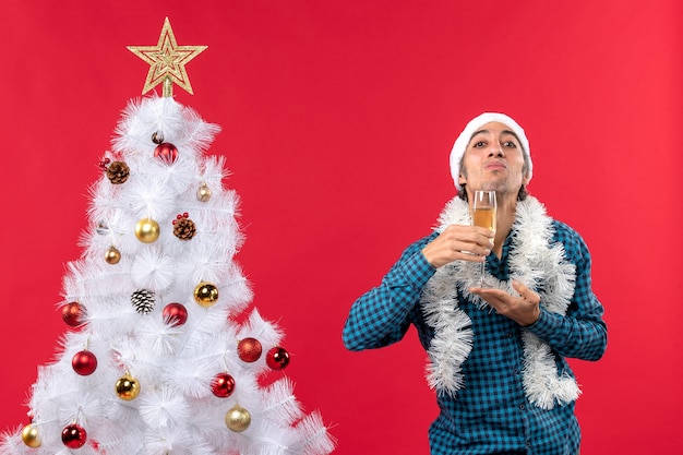 Clima natalino com jovem emocional com chapéu de papai noel em uma camisa azul listrada segurando uma taça de vinho sob o queixo perto da árvore de natal