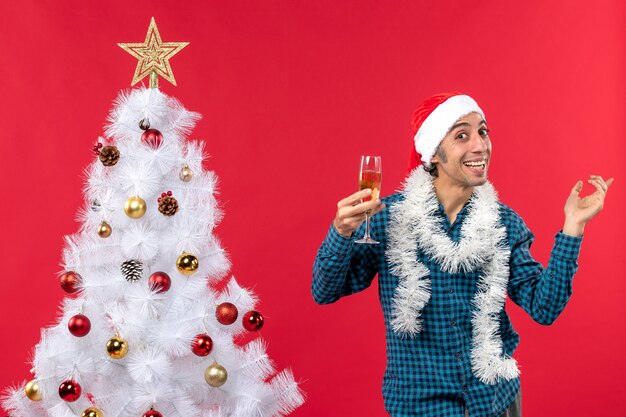 Clima natalino com jovem feliz com chapéu de papai noel em uma camisa azul listrada levantando uma taça de vinho mostrando a esquerda perto da árvore de natal