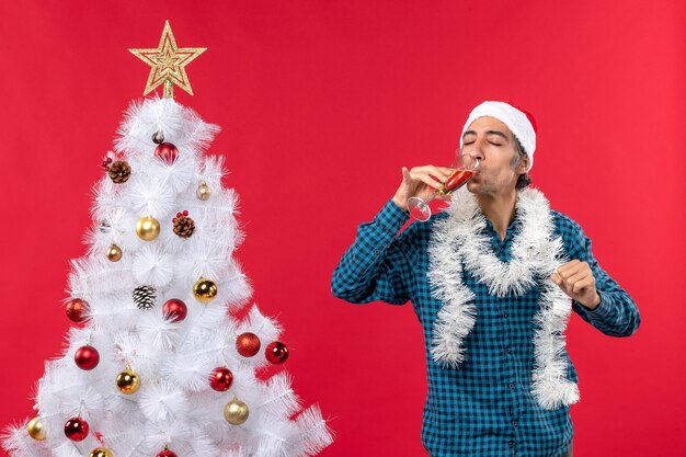 Clima natalino emocional jovem com chapéu de papai noel em uma camisa azul listrada segurando e bebendo uma taça de vinho fechando os olhos perto da árvore de natal