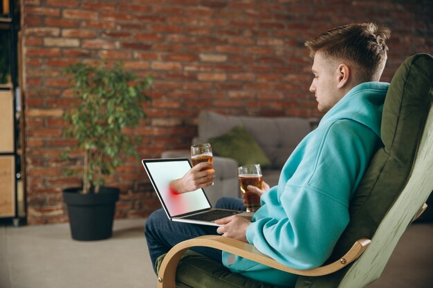 Clinking. Jovem bebendo cerveja durante uma reunião com amigos em uma videochamada virtual. Reunião online à distância, bate-papo no laptop em casa. Conceito de entretenimento e reuniões remotas seguras.